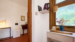 A room with a desk, a small fridge, a shelf with a kettle, a window with a view of greenery, and a hanging basket. - 'La Stanza di Campagna' with Shared Garden and Wi-Fi (Campiglia Marittima)