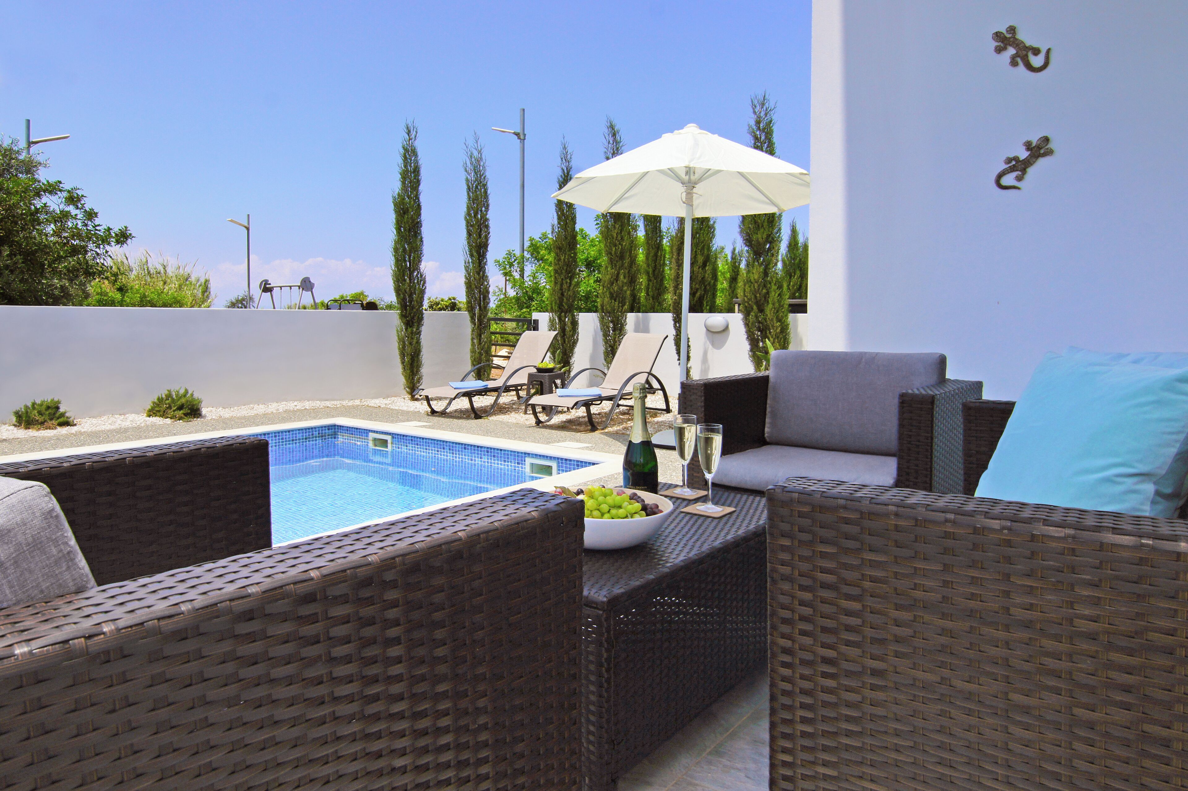 A poolside area with wicker furniture, a table with a bowl of fruit and drinks, and a white umbrella.