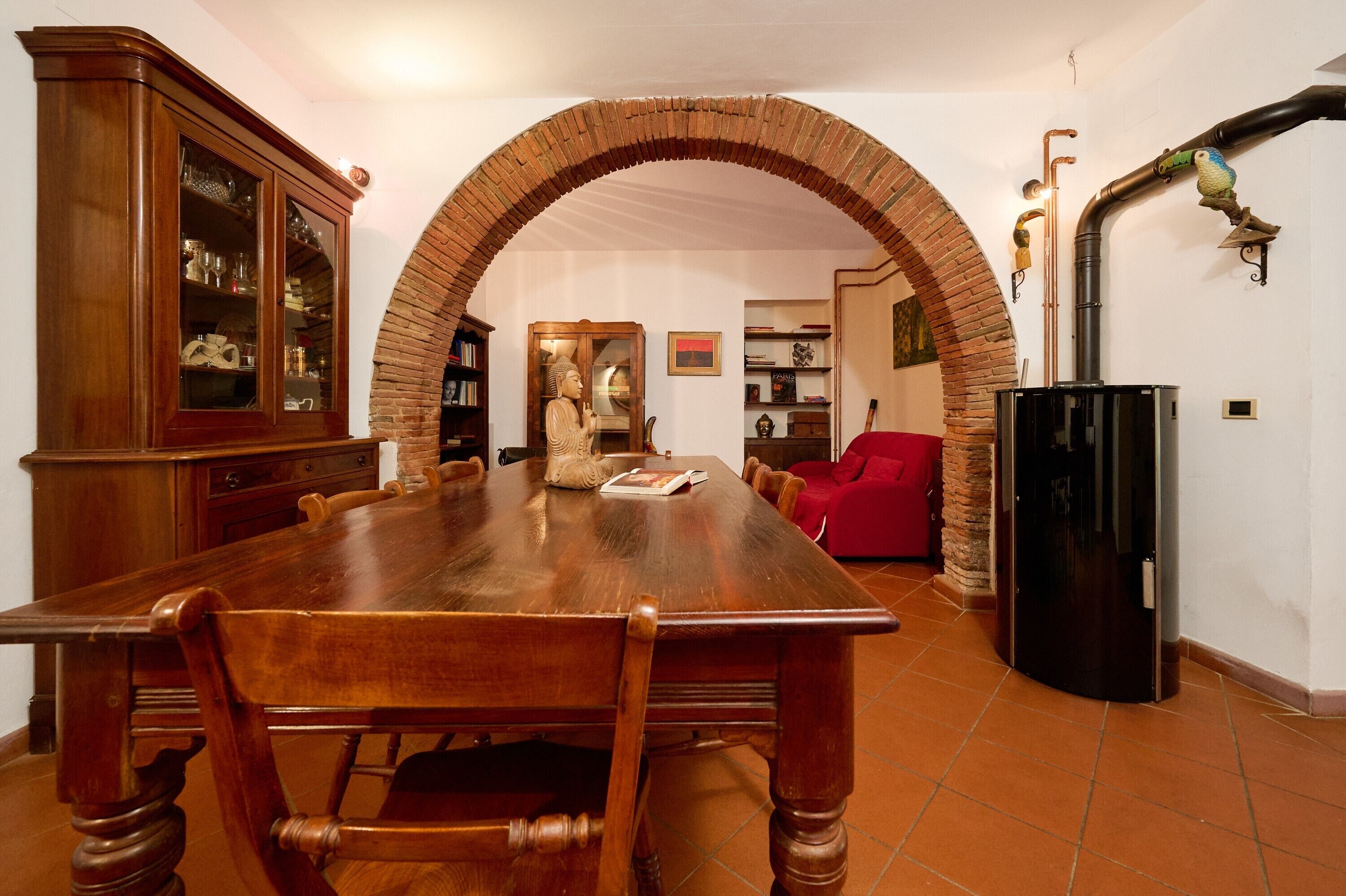 Un'area pranzo con un tavolo di legno lungo, sedie, una credenza con vetrate, una statua e un divano rosso.