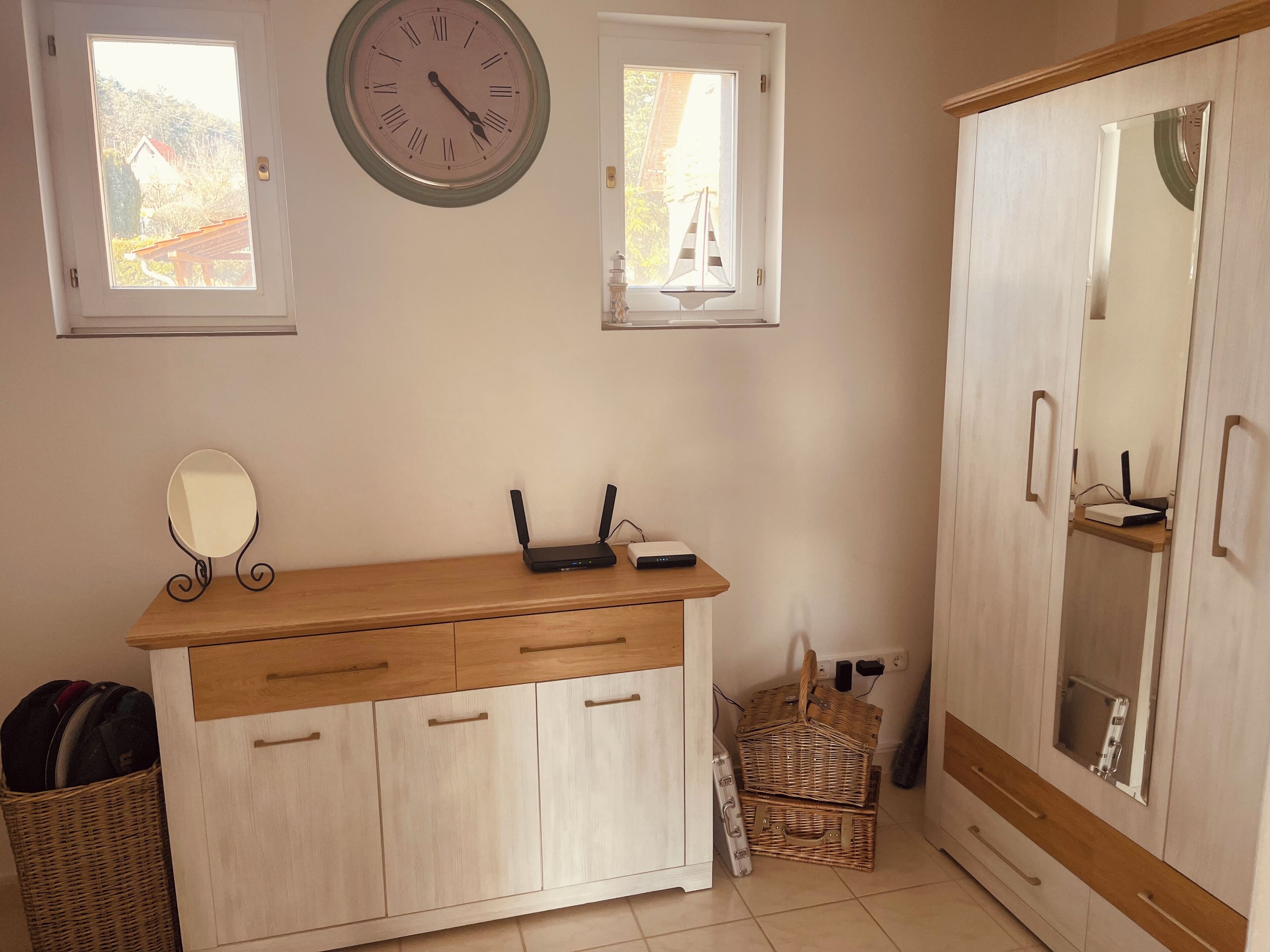 A room with a wooden cabinet, a wall clock, two windows, a mirror, a wicker basket, and a wardrobe.