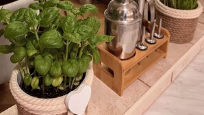 Una cocina con una maceta de albahaca, un shaker de cócteles y una bandeja de madera con utensilios de bar.