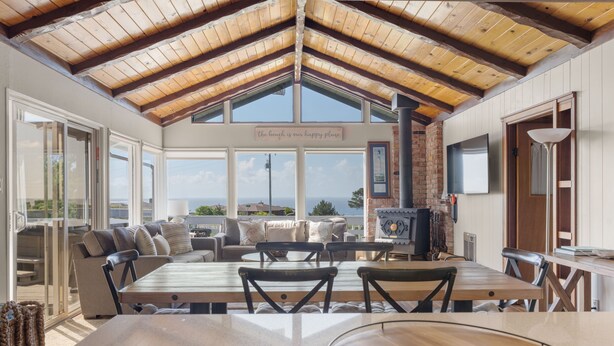A spacious room with a high wooden ceiling, large windows offering a view of the sea, a fireplace, and a dining area with a table and chairs. - Irish Beach | Ocean Views, Hot-tub, Beach Access (Manchester)