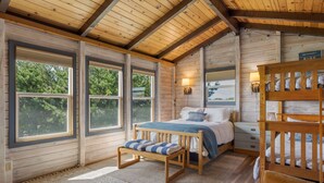 A wooden cabin bedroom with a bed, bench, and bunk beds, large windows, and a view of trees. - Irish Beach | Ocean Views, Hot-tub, Beach Access (Manchester)