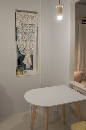 Une pièce avec une table blanche, une fenêtre ornée d’un rideau en dentelle, une plante en pot et un miroir.