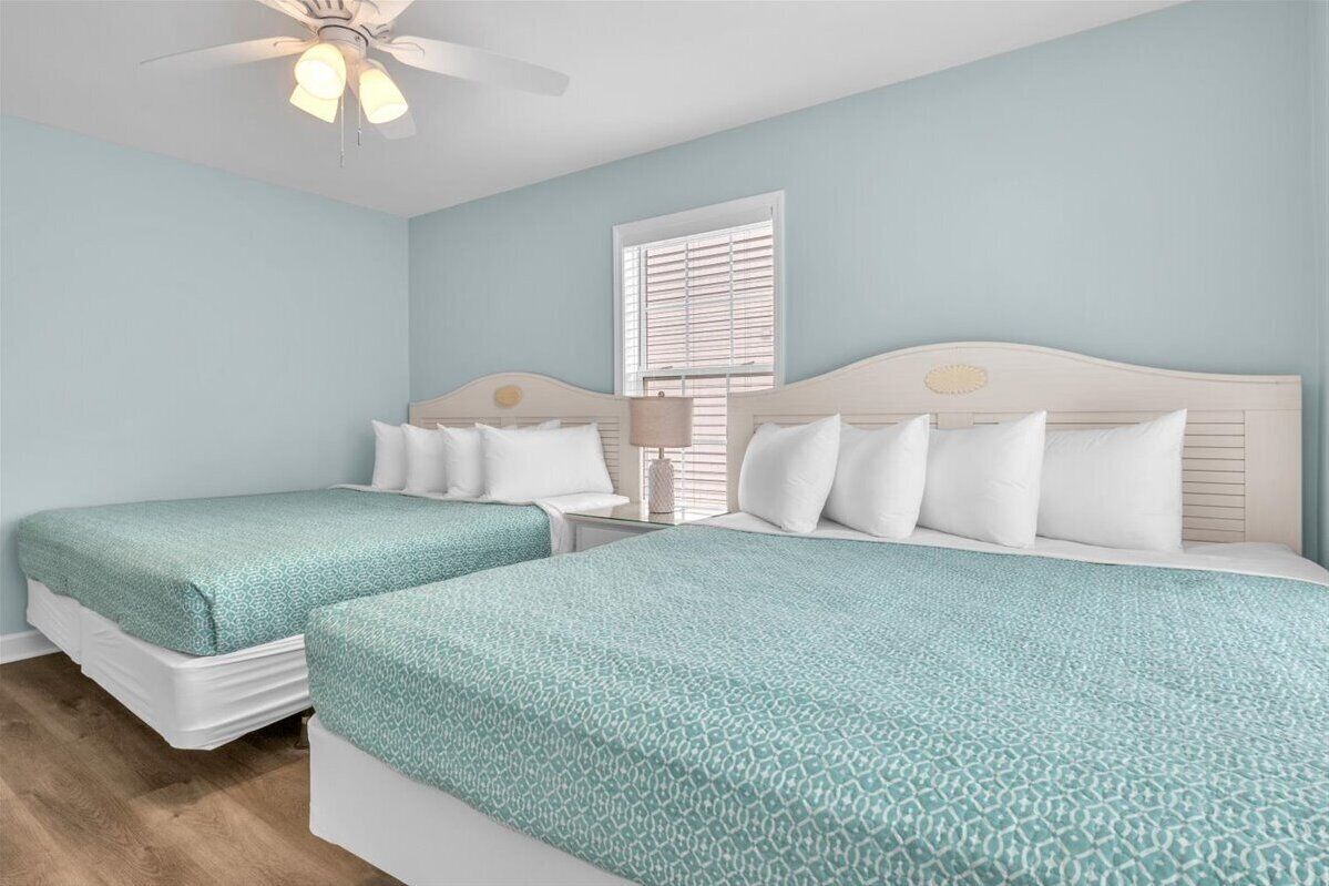 A bedroom with two beds, a ceiling fan, a window with blinds, and a bedside table with a lamp.