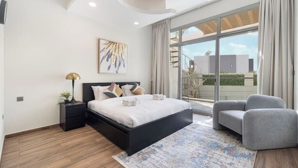 Een moderne slaapkamer met een groot bed, een nachtkastje, een lamp, een grijze fauteuil en een groot raam met uitzicht op gebouwen.