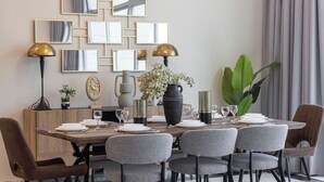 Een eetruimte met een houten tafel gedekt voor zes personen, omringd door grijze gestoffeerde stoelen. Er staan twee vloerlampen met gouden kapjes, een grote potplant en een wandspiegel-opstelling.