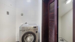 Een kleine wasruimte met een voorlader wasmachine, een klein plankje en een deur met horizontale lamellen.