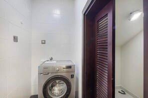 A compact laundry room with a front-loading washing machine, a small shelf, and a door with horizontal slats.