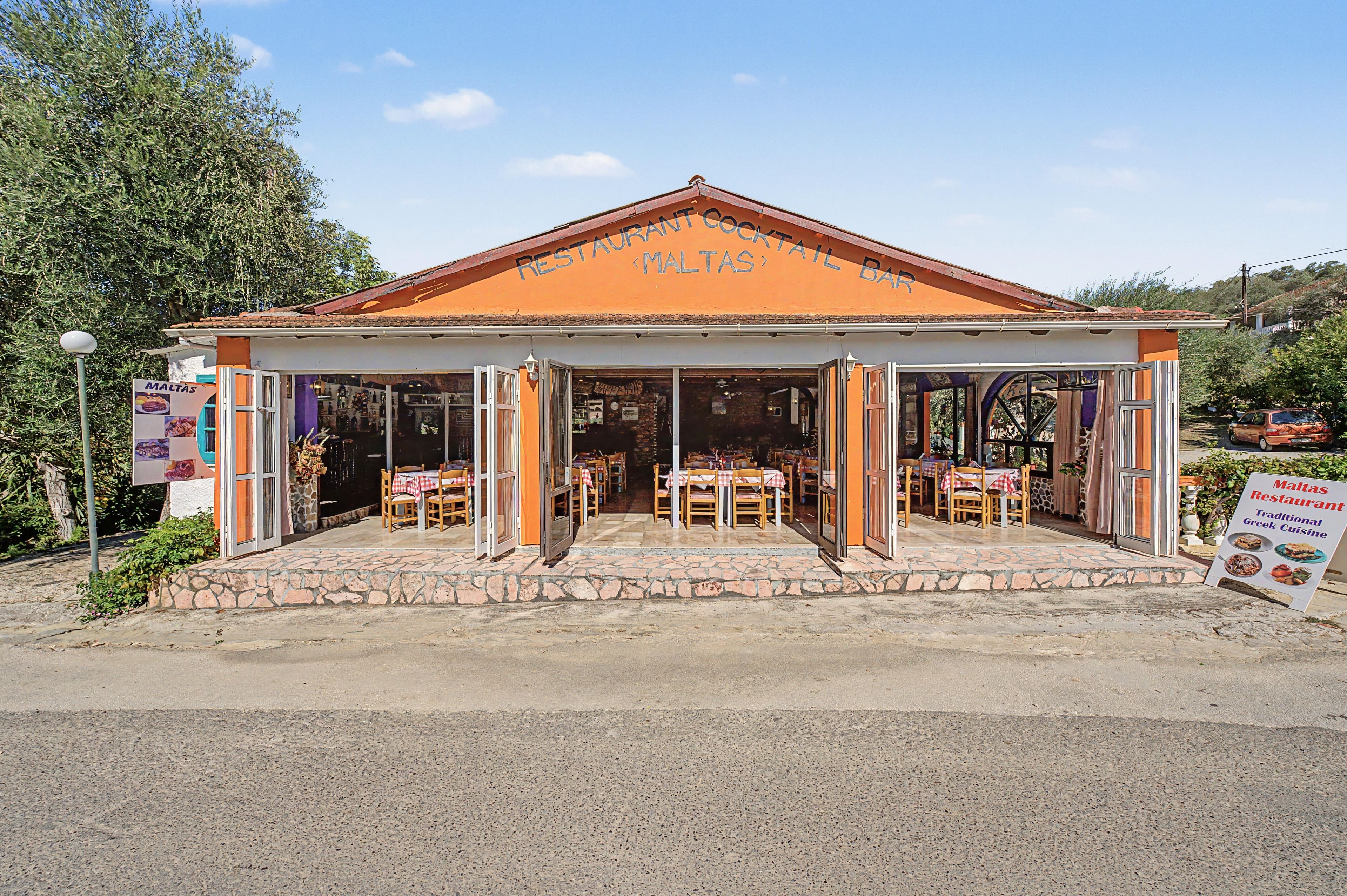 Een restaurant met een terras, een oprit met stenen bestrating en een bord dat traditionele Griekse keuken aanprijst.