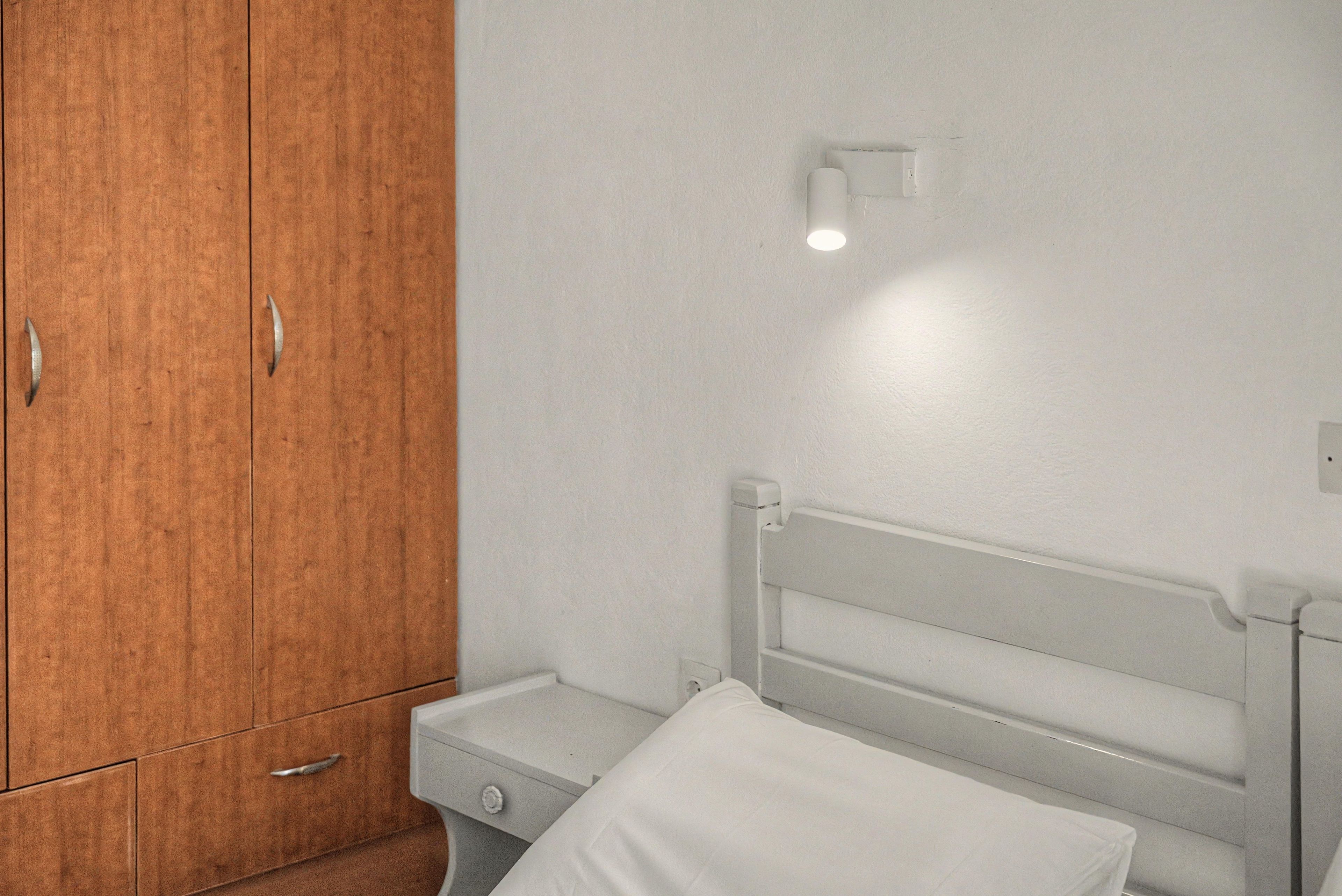 A bedroom with a wooden wardrobe, a bedside table, and a white headboard.