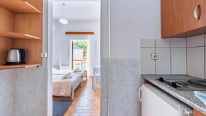 Eine moderne Küche mit Einbauherd, einer auf einem Regal stehenden Kaffeemaschine und Blick in ein Schlafzimmer mit Bett und Fenster.