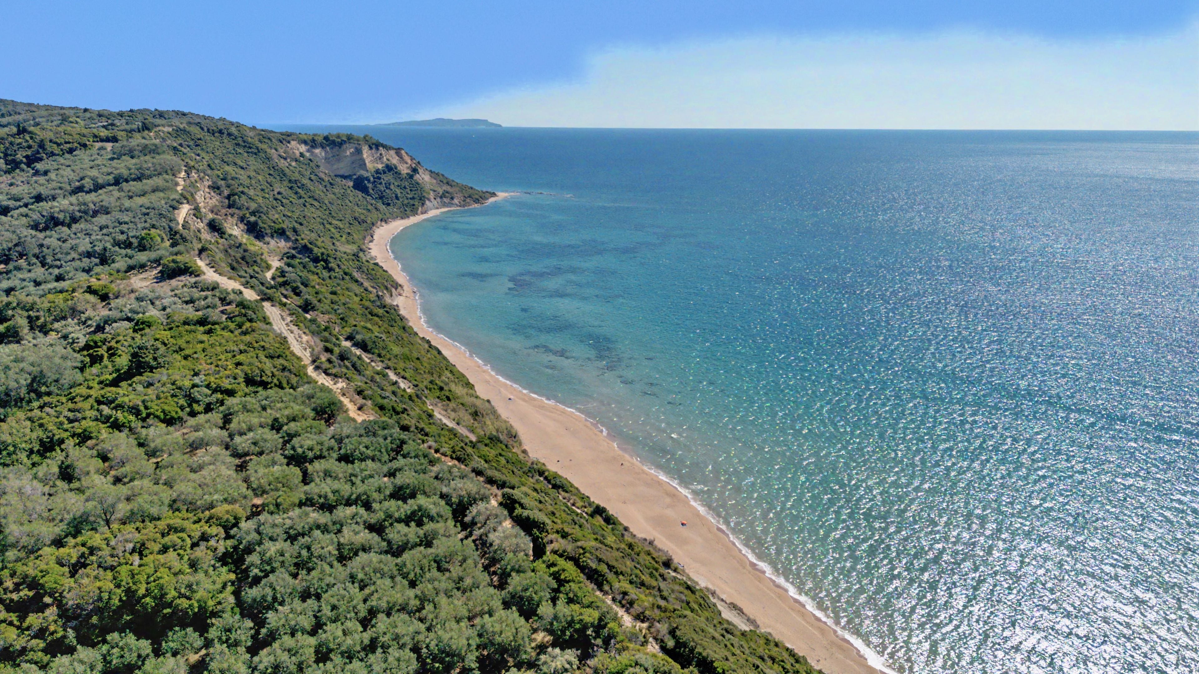 A coastal landscape with a beach, dense greenery, and a clear view of the sea.