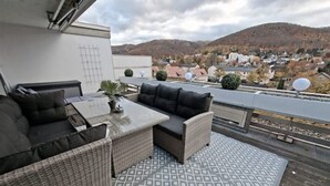 A rooftop terrace with outdoor furniture, a patterned floor, and a view of a town and hills in the background. - Bergliebe 13 - Modern vacation apartment with roof terrace & mountain views (Bad Harzburg)