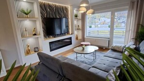 A modern living room with a grey sofa, a flat-screen TV mounted on a wall, a fireplace, and a large window offering a view of the outdoors. - Bergliebe 13 - Modern vacation apartment with roof terrace & mountain views (Bad Harzburg)
