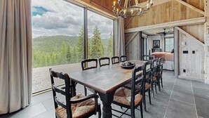 A dining area with a large wooden table and chairs, a chandelier, and a view of a forest through large windows. - Flathead Lake Barn House w/Lake & Mountain Views (Lakeside)