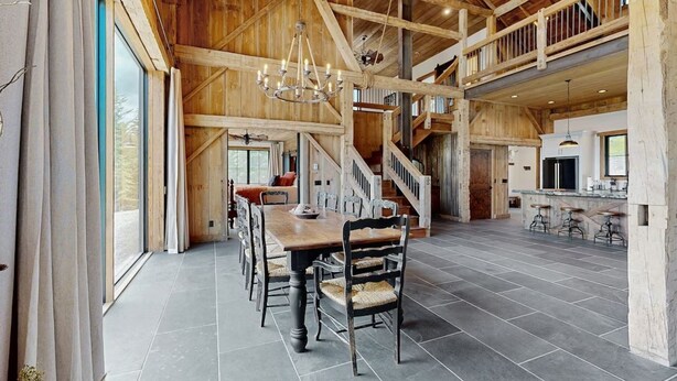 A spacious dining area with a high ceiling and wooden beams, a staircase leading to an upper level, and a kitchen area with bar stools. - Flathead Lake Barn House w/Lake & Mountain Views (Lakeside)