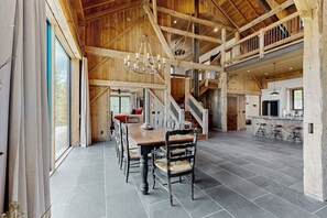 A spacious dining area with a high ceiling and wooden beams, a staircase leading to an upper level, and a kitchen area with bar stools.