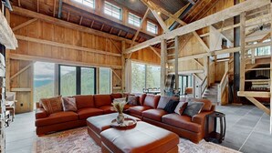 A spacious living room with a large sectional sofa, a coffee table, and a fireplace. There are large windows offering a view of the outdoors. - Flathead Lake Barn House w/Lake & Mountain Views (Lakeside)