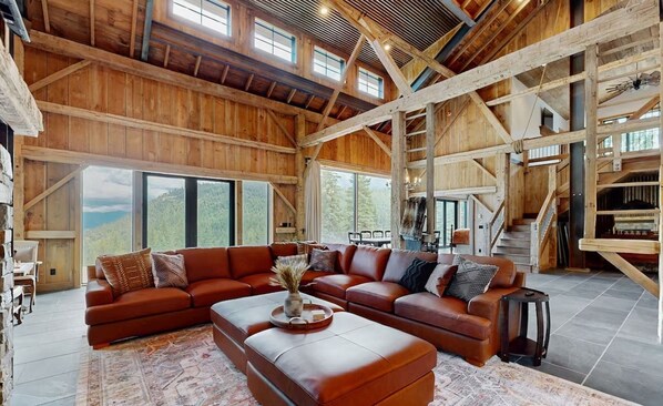 A spacious living room with a large sectional sofa, a coffee table, and a fireplace. There are large windows offering a view of the outdoors.