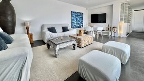A modern living room with a sofa, coffee table, ottoman, and a dining area with chairs and a TV. - Villa Sol in Canyelles with private pool and Wi-Fi (Roses)