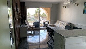 Ein modernes Wohnzimmer mit Sofa, Sesseln und Essbereich. Ein Balkon bietet Blick auf Palmen und Gebäude.