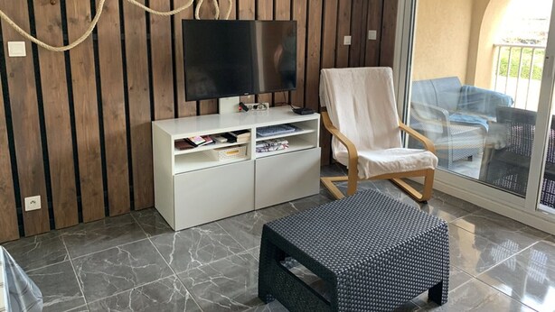 Ein modernes Wohnzimmer mit einer Holzvertäfelung, einem Flachbildfernseher, einem weißen Sessel, einem kleinen schwarzen Hocker und einer Glastür zu einem Außenbereich.