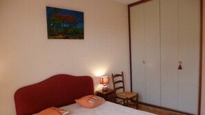 A bedroom with a bed, pillows, a nightstand with a lamp, a chair, a wardrobe, and a painting on the wall. - Charming T2 near Thau Lagoon - 2 guests (Balaruc-les-Bains)