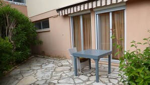 A stone-floored patio with a table and chair, a window with curtains, and a small plant. - Charming T2 near Thau Lagoon - 2 guests (Balaruc-les-Bains)