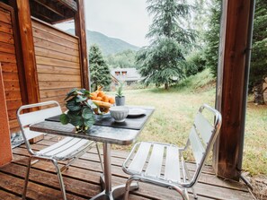 Une cabane en bois avec une table et deux chaises sur une terrasse, d’où l’on peut admirer une zone boisée avec des montagnes en arrière-plan.
