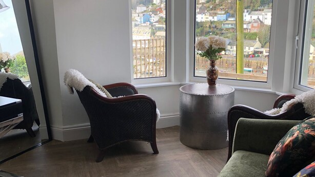 En stue med en grøn sofa, en fletstol og et rundt bord med en blomstervase. Store vinduer giver udsigt over en bakke med huse.