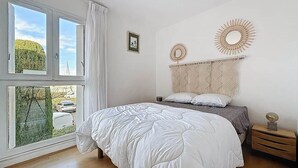 A bedroom with a large window, a bed with white bedding, a wooden nightstand, and a wall-mounted mirror. - Marina Saphirre-Port Ulysse -P2 (Le Grau-du-Roi)