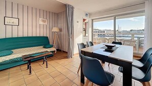 A living room with a sofa, a dining table, and chairs. There is a window with a view of a marina. - Marina Saphirre-Port Ulysse -P2 (Le Grau-du-Roi)