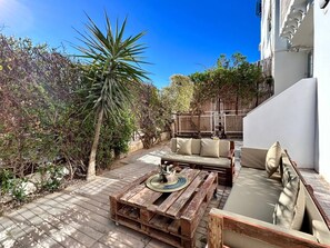 An outdoor patio with a wooden table and a sofa, surrounded by greenery and a white building. - Sunny Blue Apartment in la Marsa (La Marsa)