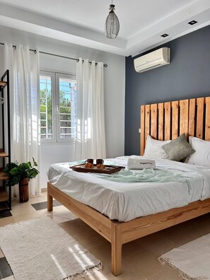 A bedroom with a wooden bed, white bedding, a SuperHost sign, a potted plant, and a view of greenery outside. - Sunny Blue Apartment in la Marsa (La Marsa)
