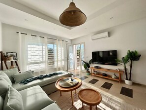 A modern living room with a sofa, two small tables, a TV, and a large window with curtains. - Sunny Blue Apartment in la Marsa (La Marsa)