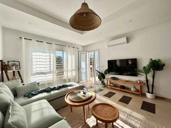 A modern living room with a sofa, two small tables, a TV, and a large window with curtains. - Sunny Blue Apartment in la Marsa (La Marsa)