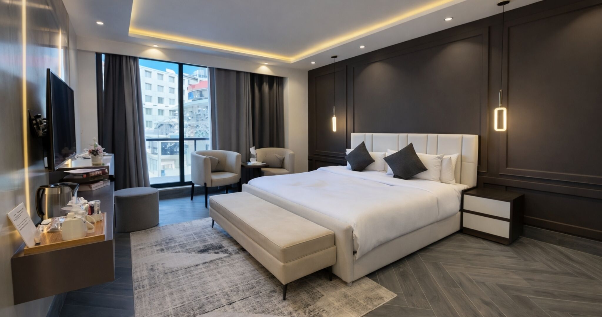 A modern hotel room with a large bed, bedside tables, a seating area, and a view of the city.