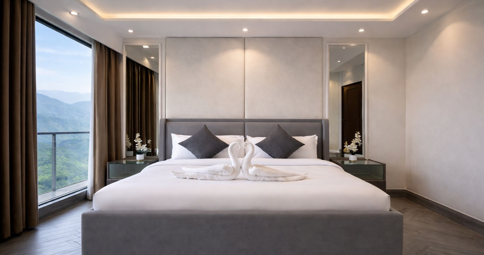 A modern hotel room with a large bed, two nightstands, a mirror, and a view of the mountains.
