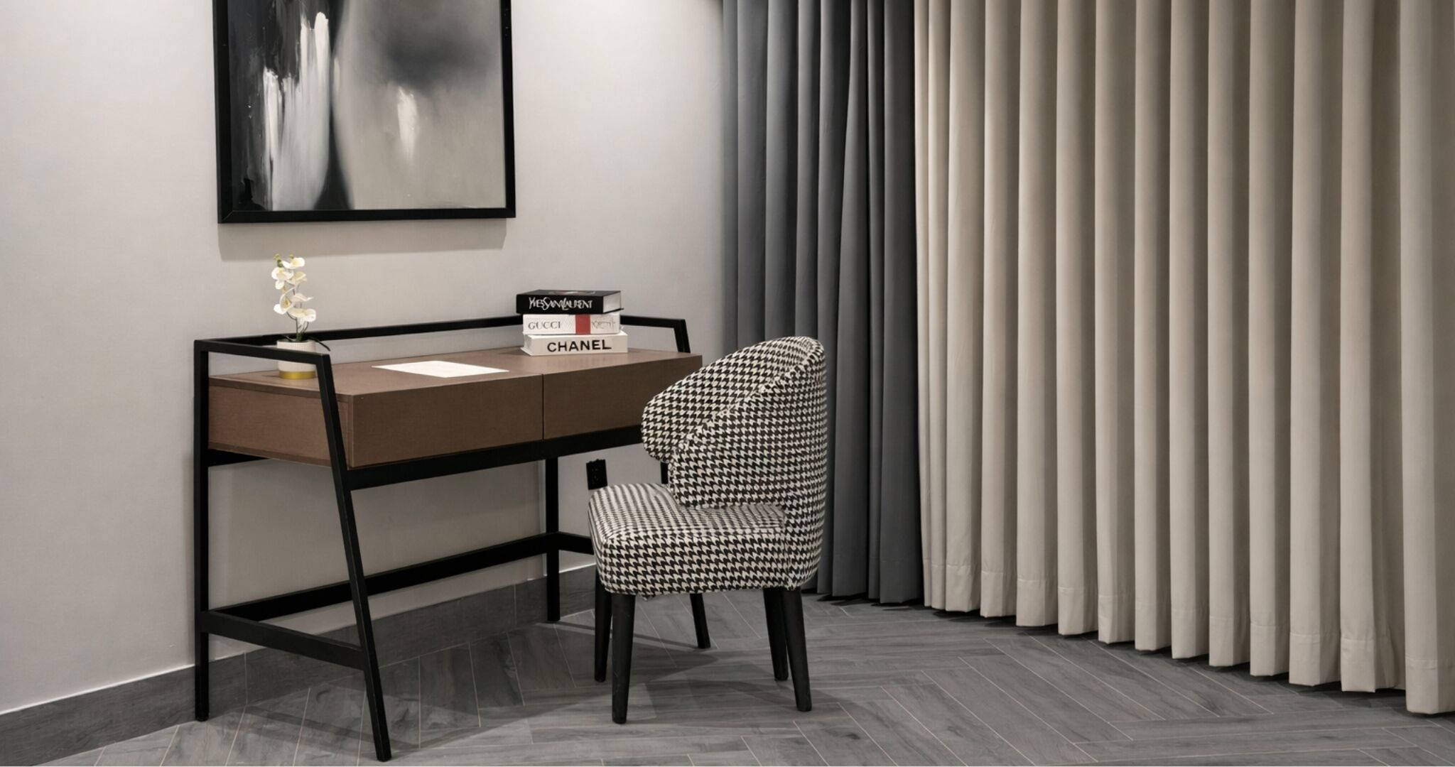 A modern office space with a desk, a houndstooth patterned chair, a framed abstract painting, and a set of books.