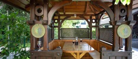 A wooden gazebo with a table and two clocks, surrounded by greenery.