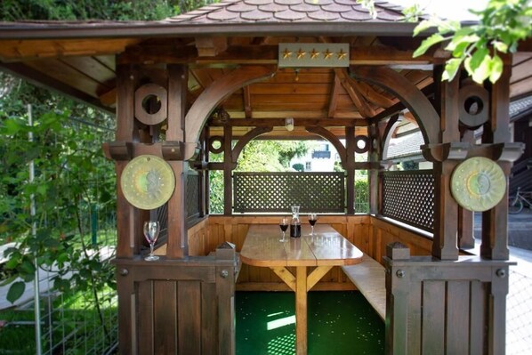 A wooden gazebo with a table and two clocks, surrounded by greenery.