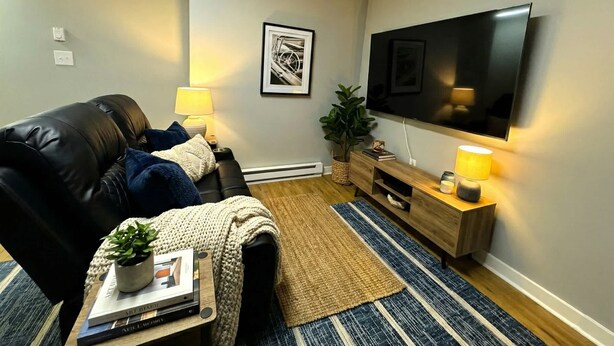 En stue med en sort lædersofa, et fladskærms-tv, et sofabord med bøger og en plante, en lampe og et billede i ramme på væggen.