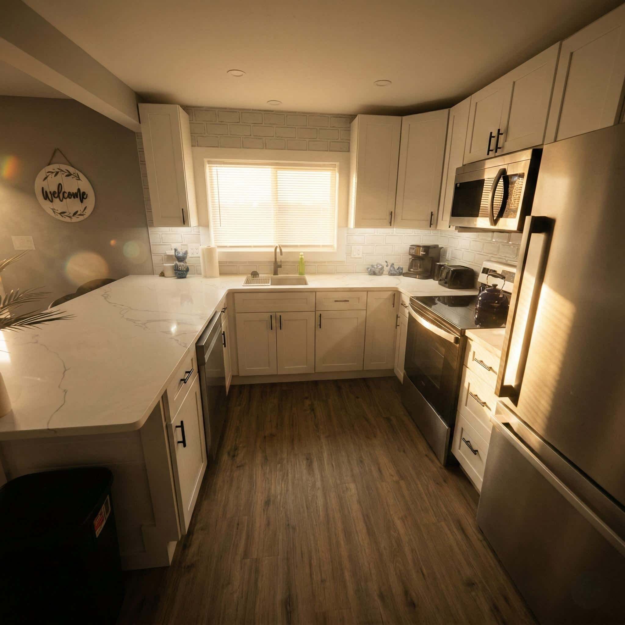 A modern kitchen with white cabinets, a marble countertop, stainless steel appliances, and a window with blinds.