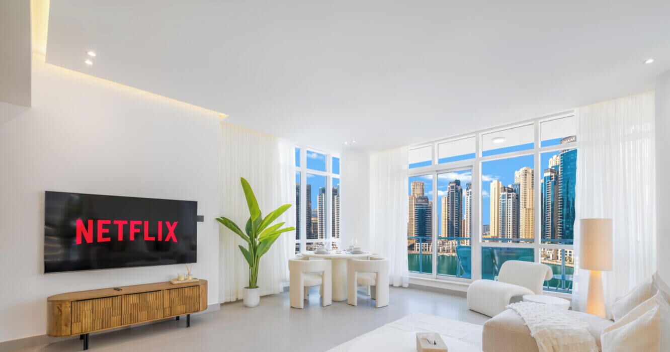 A modern living room with a large TV displaying "NETFLIX," a dining area with a table and chairs, and a city view through large windows.
