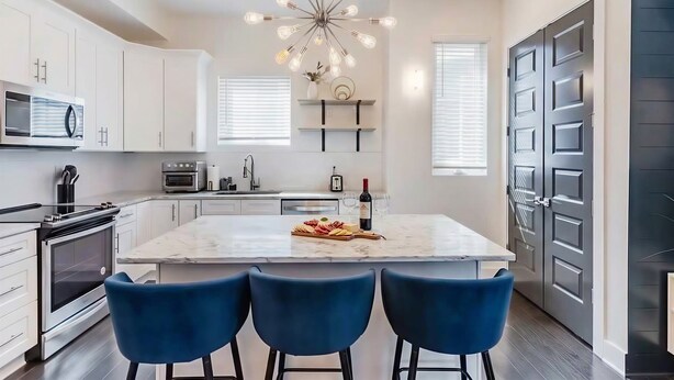 A modern kitchen with white cabinets, a central island with blue bar stools, a stainless steel oven, and a unique light fixture. - Rooftop Views Patio Lounge Near Downtown Nashville (Nashville)