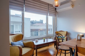 A modern living room with a wooden dining table, two sofas, and a large window with blinds.