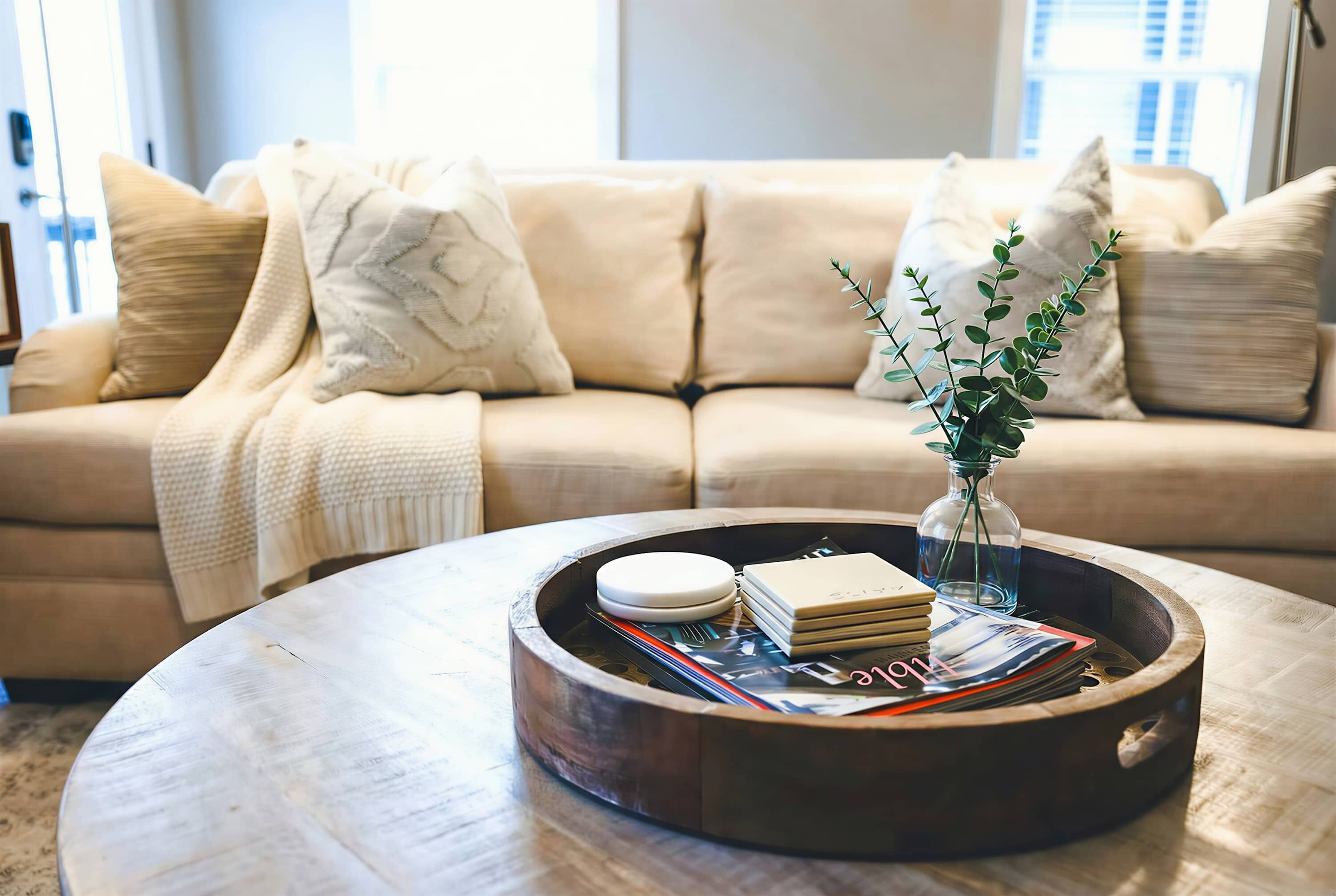 A round wooden coffee table with a tray containing books, a vase with a plant, and a glass of water.