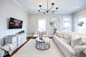 A living room with a white sofa, a round coffee table, a chandelier, a flat-screen TV, and a large mirror. - Private Pool Game Room Compound Minutes to Broadway (Nashville)