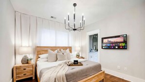 Un dormitorio moderno con cama de madera, mesitas de noche, una araña y un televisor montado que muestra un paisaje urbano.